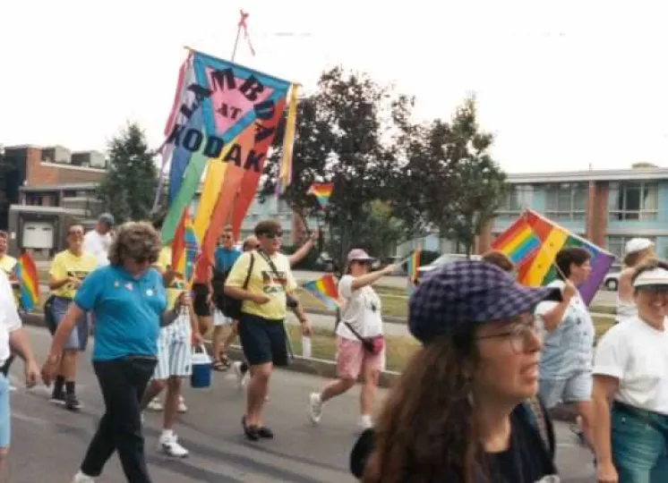 People march outdoors in a pride parade, carrying rainbow flags and a large GEM Lambda Kodak banner. Participants wear casual clothes, and buildings and trees are visible in the background. Even before they formally acquired official recognition at Kodak, Lambda members met as a support group and participated in off-campus events such as this mid-1990s Pride Parade in Rochester, NY.
