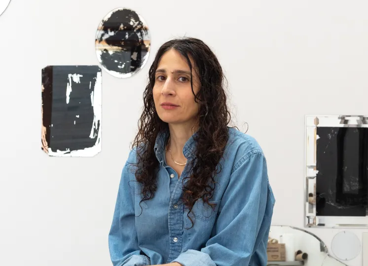 A woman with long curly hair in a denim shirt and jeans sits on a stool in an art studio, surrounded by abstract mirrored art pieces on the walls—each work inviting viewers to question fantasy and perception. Lecture event Thursday, 6 p.m. in Dryden Theatre at the George Eastman Museum.