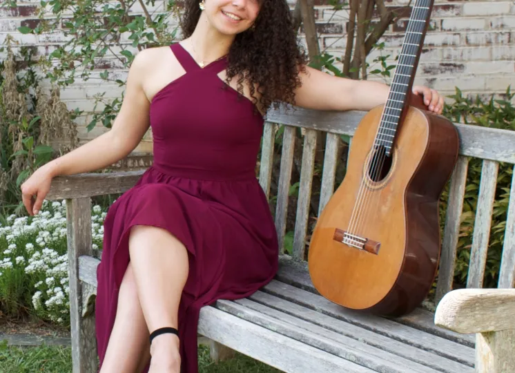 Maya Loncar, guitarist, in a long dress and sitting on a bench with her guitar outside