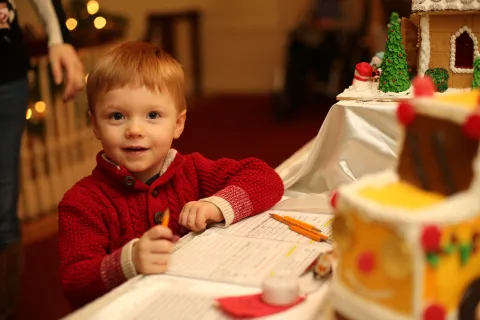 Little boy in sweater looking directly at the viewer with a pencil in his hand, a list at the table, and gingerbread houses in the background