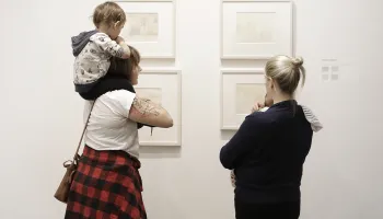 Two adults each holding a child looking at framed art on a wall