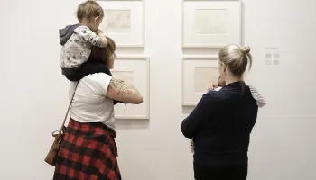 Two adults each holding a child looking at framed art on a wall