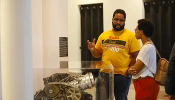 A Black man in a Kodak sweater with glasses and a Black woman with short hair and glasses having a conversation in front of a camera on display