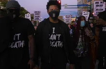 Two people at a march wearing t-shirts with the words I Can't Breathe, among a crowd of other people holding several signs with the words "Jail All Racist Killer Cops" 
