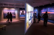 Exhibition gallery featuring large color photographs and a projection screen with five adults looking at art throughout the space