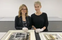Two women stand side by side facing the camera behind a table on which there are stacks of photographs