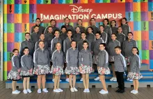 32 young adults and children stand in 4 rows facing the camera, in front of a banner that reads Disney Imagination Campus