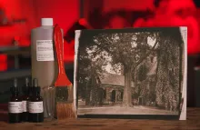 Display of photography materials which includes potassium 1000 ml bottle, a large painting brush, a small measuring vessel, and three contains of solution. Next to the materials is a vintage photo of a large tree in front of an old stone church-like structure. In the background is a blurred image of a photographic darkroom.  