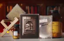 Display of photographic materials including wooden drying rack, bottle of silver nitrate, bag of sodium chloride, bottle of ammonium chloride, and salt print photograph of a skull and books