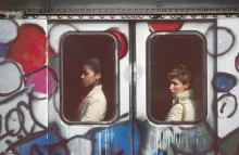 Graffiti-covered subway car from the outside, with a person visible in each of the two windows