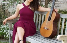 Maya Loncar, guitarist, in a long dress and sitting on a bench with her guitar outside