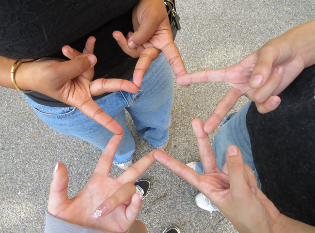 Four people form a star shape by touching their fingers together while standing in a circle on a paved surface—a perfect moment of connection and creativity captured by Click & Tell, a summer photography club for teens that celebrates curiosity and imagination as we look ahead to 2027.
