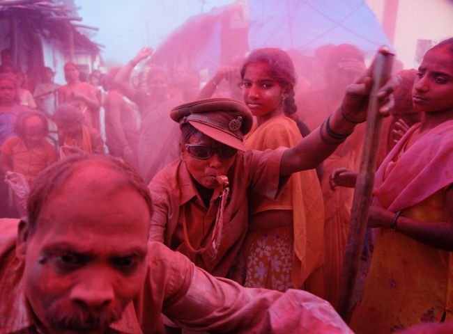 Crowd of people outdoors with one central figure blowing a whistle and another young woman looking into the camera, with magenta powder (gulal?) covering everything in view