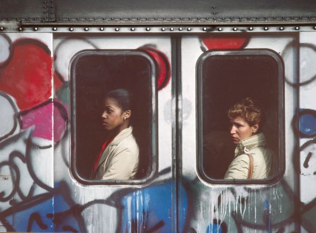 Graffiti-covered subway car from the outside, with a person visible in each of the two windows