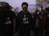 Two people at a march wearing t-shirts with the words I Can't Breathe, among a crowd of other people holding several signs with the words "Jail All Racist Killer Cops" 
