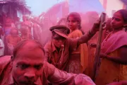 Crowd of people outdoors with one central figure blowing a whistle and another young woman looking into the camera, with magenta powder (gulal?) covering everything in view