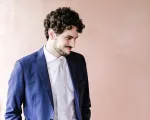 Young man in a suit with curly hair and light facial hair slightly looking down and smiling. 