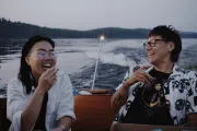 Two people laugh joyfully while sitting on a boat during a calm evening ride, with water and a forested shoreline in the background.