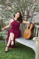 Maya Loncar, guitarist, in a long dress and sitting on a bench with her guitar outside